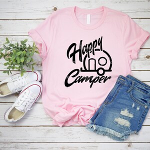 Op de afbeelding: Een roze T-shirt met zwarte tekst die "Happy Camper" zegt en een zwart silhouet van een caravan.