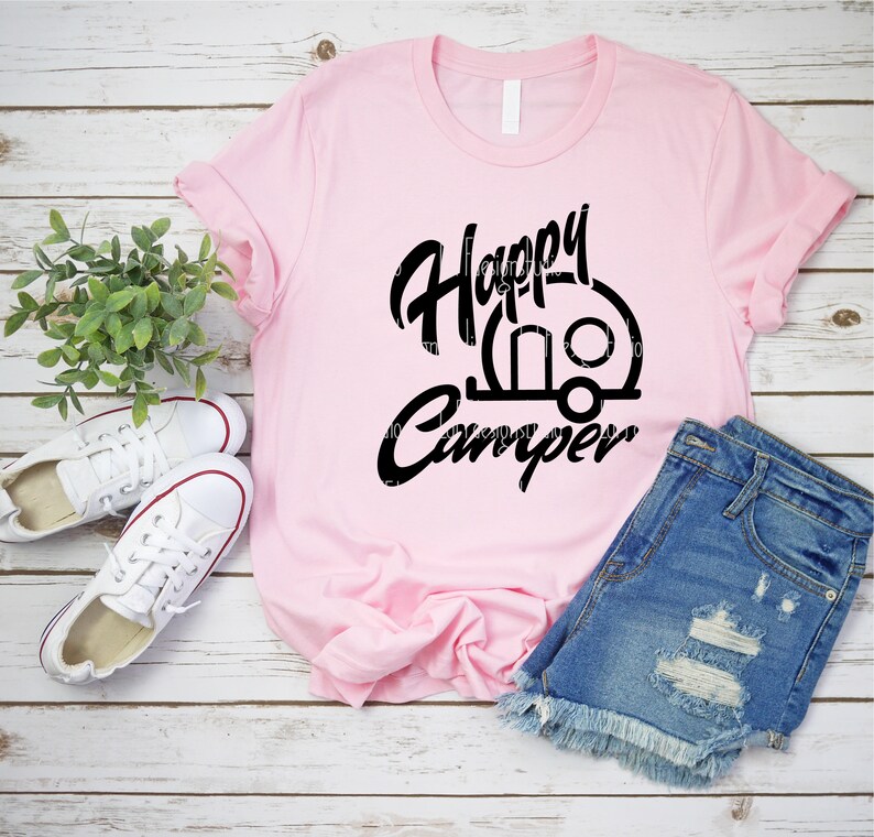 Op de afbeelding: Een roze T-shirt met zwarte tekst die "Happy Camper" zegt en een zwart silhouet van een caravan.