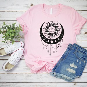 May include: A pink t-shirt with a black sun and moon design. The design features a sun with a face and a crescent moon with three smaller moons hanging from it. The design is surrounded by stars and diamonds.