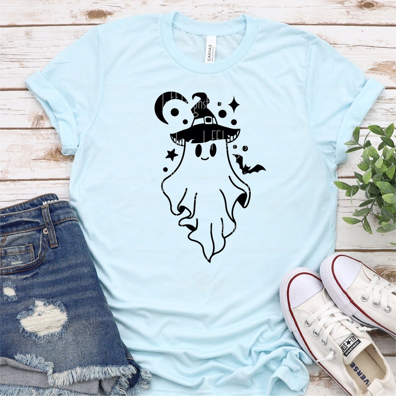 May include: A light blue t-shirt with a black and white graphic of a ghost wearing a witch hat. The ghost has a smiling face and is surrounded by stars and crescent moons.