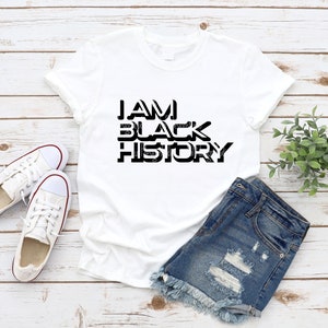 May include: White t-shirt with black text that reads "I AM BLACK HISTORY". The shirt is laying on a wooden surface with a pair of white sneakers and denim shorts.