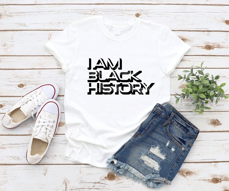 May include: White t-shirt with black text that reads "I AM BLACK HISTORY". The shirt is laying on a wooden surface with a pair of white sneakers and denim shorts.