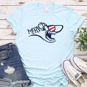 May include: A light blue t-shirt with a shark graphic wearing red, white, and blue sunglasses. The shark is patterned with stars and stripes and the word "MERICA" is written in a curved font.