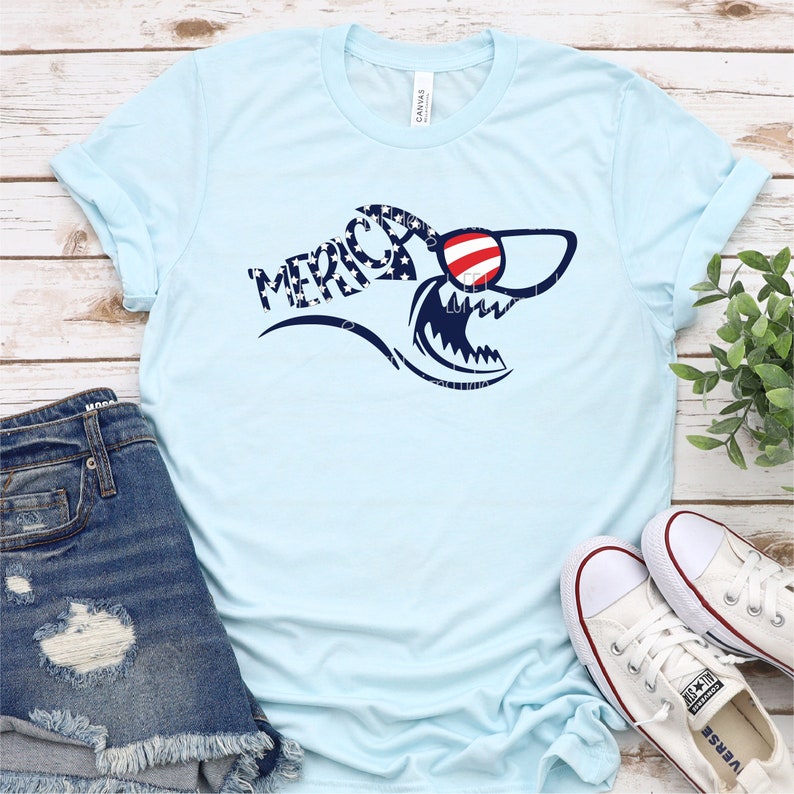 May include: A light blue t-shirt with a shark graphic wearing red, white, and blue sunglasses. The shark is patterned with stars and stripes and the word "MERICA" is written in a curved font.