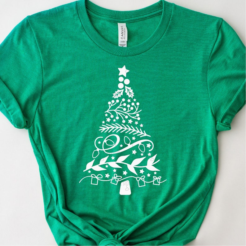 Op de afbeelding: Een groen T-shirt met een witte omtrek van een kerstboom versierd met kerstballen, sterren en cadeautjes.