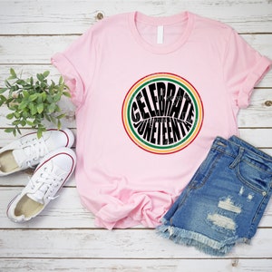 K&ouml;nnte beinhalten: Pinkes T-Shirt mit einem schwarzen Kreis-Motiv, auf dem in einer Retro-Schriftart "Celebrate Juneteenth" steht. Der Kreis ist von roten, gelben und gr&uuml;nen Streifen umgeben.