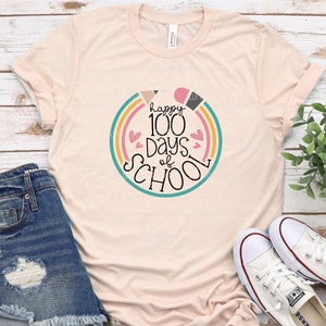May include: A light pink t-shirt with a graphic design that says "happy 100 days of school" in a colorful circle with rainbow stripes and hearts.