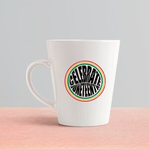 K&ouml;nnte beinhalten: Eine wei&szlig;e Keramiktasse mit einem schwarzen und gr&uuml;nen Kreisdesign, auf dem "Celebrate Juneteenth" steht.