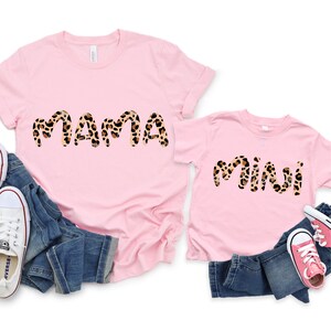 K&ouml;nnte beinhalten: Zwei rosa T-Shirts mit den Worten "mama" und "mini" in einem Leopardenmuster. Die Shirts liegen auf einem blauen Jeansstoff mit rosa und wei&szlig;en Turnschuhen.