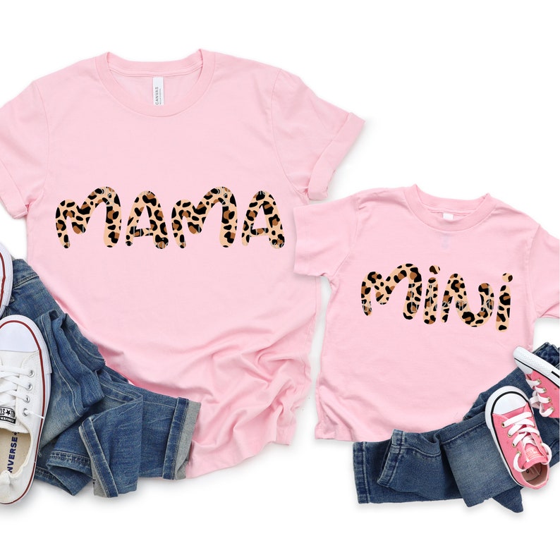 K&ouml;nnte beinhalten: Zwei rosa T-Shirts mit den Worten "mama" und "mini" in einem Leopardenmuster. Die Shirts liegen auf einem blauen Jeansstoff mit rosa und wei&szlig;en Turnschuhen.