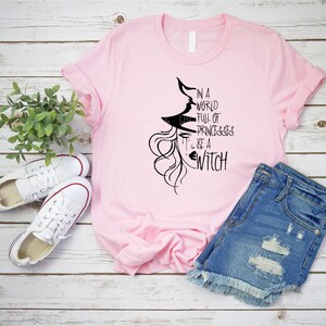 Peut inclure: T-shirt rose avec un graphique noir d'une sorci&egrave;re avec le texte "Dans un monde plein de princesses... sois une sorci&egrave;re".