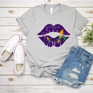 May include: A gray t-shirt with a purple lips design featuring green and gold fleur-de-lis. The shirt is laid out on a wood surface with a pair of white sneakers and denim shorts.
