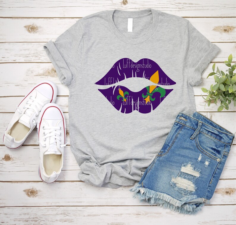 May include: A gray t-shirt with a purple lips design featuring green and gold fleur-de-lis. The shirt is laid out on a wood surface with a pair of white sneakers and denim shorts.