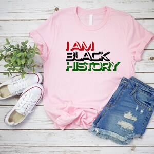 May include: A pink t-shirt with the text "I AM BLACK HISTORY" in black, red, and green letters. The shirt is laid out on a wooden surface with a pair of white sneakers and blue denim shorts.