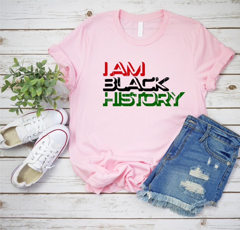 May include: A pink t-shirt with the text "I AM BLACK HISTORY" in black, red, and green letters. The shirt is laid out on a wooden surface with a pair of white sneakers and blue denim shorts.