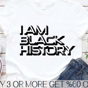 Puede incluir: Una camiseta blanca con texto negro que dice "I AM BLACK HISTORY". El texto está en negrita, con una fuente de bloque. La camiseta está sobre una superficie de madera con un par de pantalones vaqueros y una planta en el fondo.