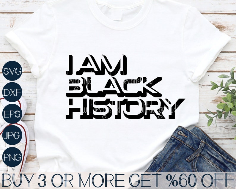 May include: A white t-shirt with black text that reads "I AM BLACK HISTORY". The text is in a bold, blocky font. The shirt is on a wooden surface with a pair of jeans and a plant in the background.