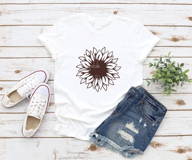 Puede incluir: Una camiseta blanca con un dise&ntilde;o gr&aacute;fico de girasol marr&oacute;n. La camiseta est&aacute; colocada sobre una superficie de madera con un par de zapatillas blancas y pantalones cortos de mezclilla.