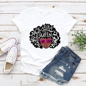 May include: White t-shirt with a black and white graphic of a woman with an afro. The afro is made up of the words "Magic", "Queen", "Strong", "Love", and "Black". The woman is wearing pink sunglasses.