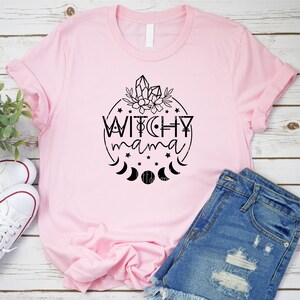 May include: Pink t-shirt with a black graphic design of a circle with the text "Witchy mama" inside. The circle is surrounded by stars, moons, and crystals.