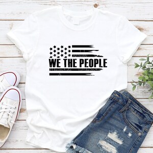 Op de afbeelding: Wit T-shirt met een verweerd Amerikaans vlagontwerp in zwart en de tekst "WE THE PEOPLE". Het shirt wordt gecombineerd met een spijkerbroek en witte sneakers, wat zorgt voor een casual, patriottische uitstraling.