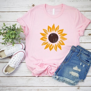 May include: A pink t-shirt with a yellow sunflower graphic. The sunflower has a brown center and is on a pink t-shirt. The t-shirt is laying on a white wooden surface.