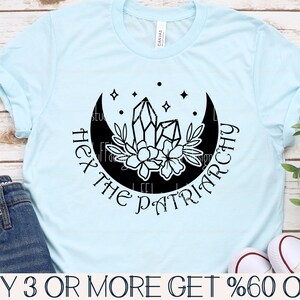 May include: A light blue t-shirt with a black graphic design of a crescent moon with crystals and flowers. The text "Hex the Patriarchy" is written around the moon.