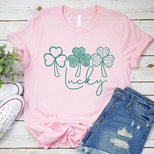 May include: A pink t-shirt featuring three green shamrock designs and the word "lucky" written in green cursive.