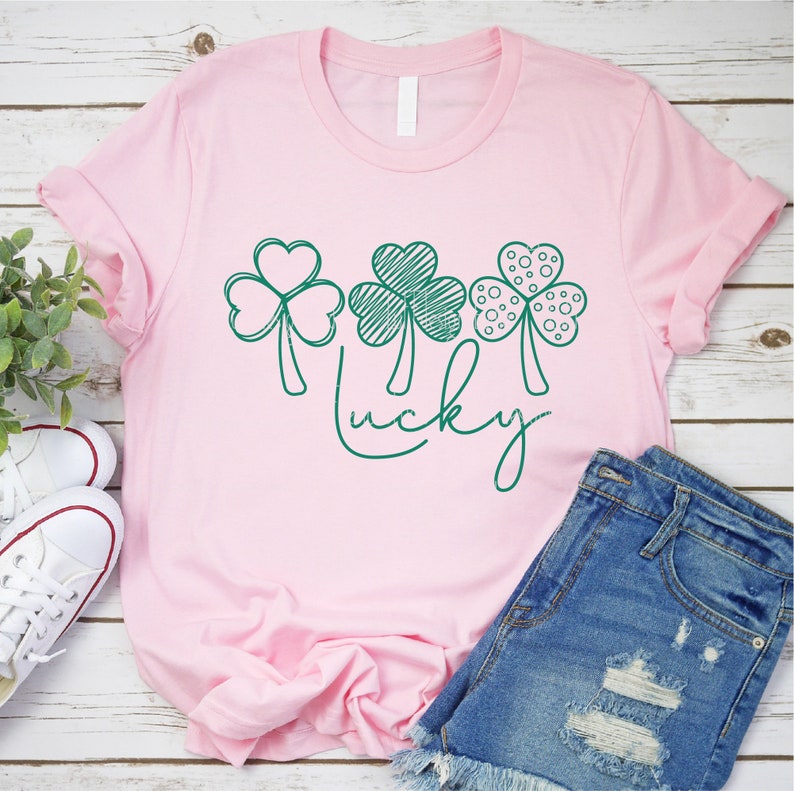 May include: A pink t-shirt featuring three green shamrock designs and the word "lucky" written in green cursive.