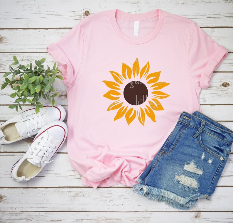 Puede incluir: Una camiseta rosa con un gr&aacute;fico de girasol amarillo. El girasol tiene un centro marr&oacute;n y est&aacute; centrado en la camiseta. La camiseta est&aacute; sobre una superficie de madera blanca.