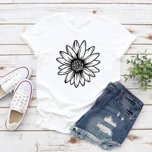May include: A white t-shirt with a black outline of a daisy flower on the front. The t-shirt is laying on a wooden surface with a pair of white sneakers and denim shorts.