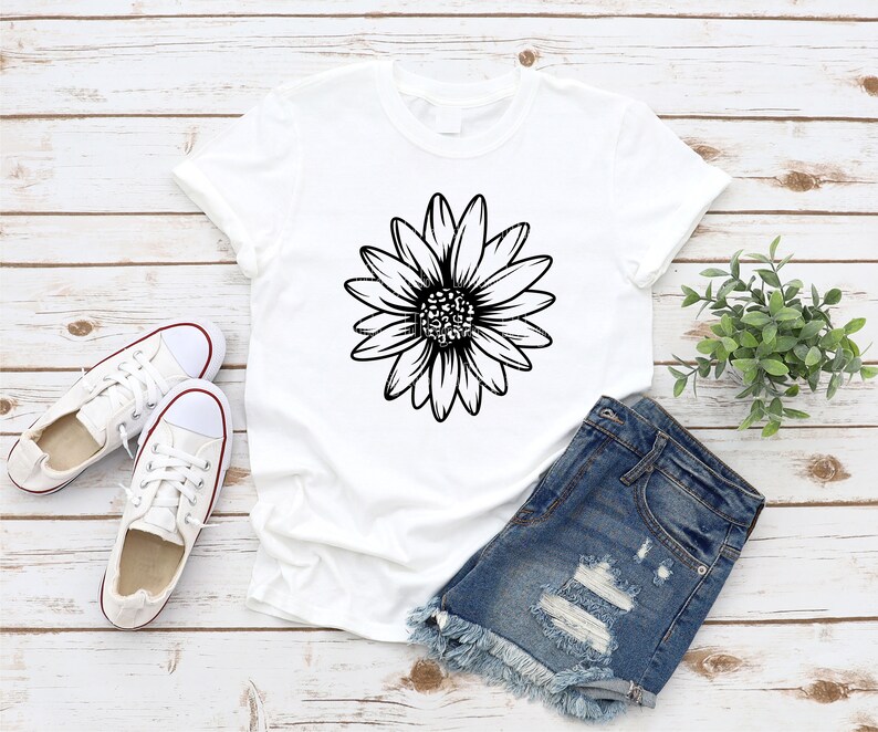 May include: A white t-shirt with a black outline of a daisy flower on the front. The t-shirt is laying on a wooden surface with a pair of white sneakers and denim shorts.