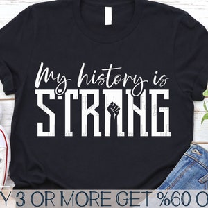May include: Black t-shirt with white text that reads "My history is strong" with a raised fist graphic. The text is in a bold, blocky font.