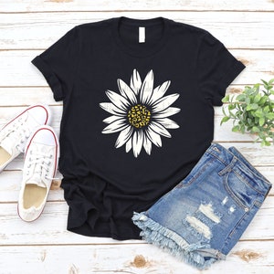 Op de afbeelding: Een zwart T-shirt met een witte madeliefje-print met een gele kern met een luipaardprint. Het shirt ligt op een wit houten oppervlak met een paar witte canvas schoenen en een blauwe denim short.