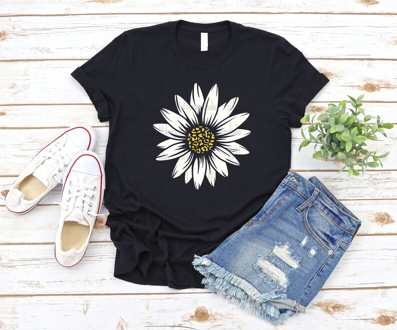 Op de afbeelding: Een zwart T-shirt met een witte madeliefje-print met een gele kern met een luipaardprint. Het shirt ligt op een wit houten oppervlak met een paar witte canvas schoenen en een blauwe denim short.