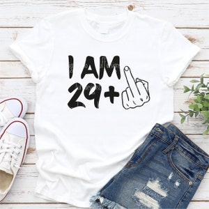 May include: White t-shirt with black text that reads "I AM 29+" and a black silhouette of a middle finger.