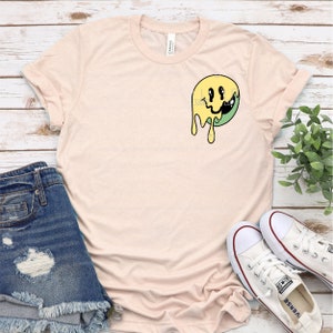 May include: A light pink t-shirt with a yellow and green dripping smiley face graphic.