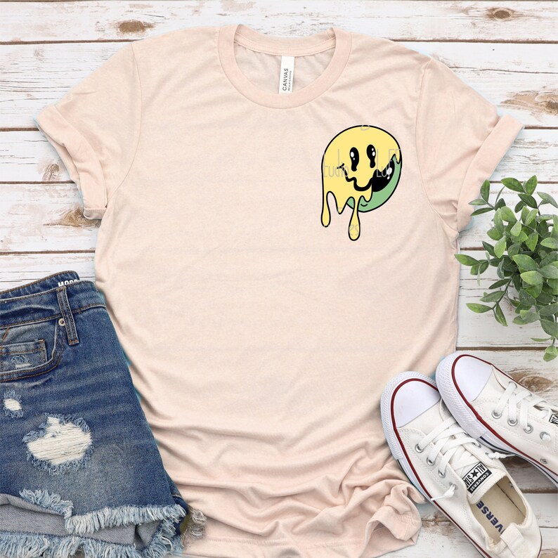 May include: A light pink t-shirt with a yellow and green dripping smiley face graphic.