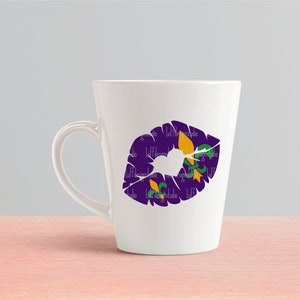 May include: White ceramic mug with a purple and gold lip print featuring a fleur-de-lis design. The mug has a handle on the right side.