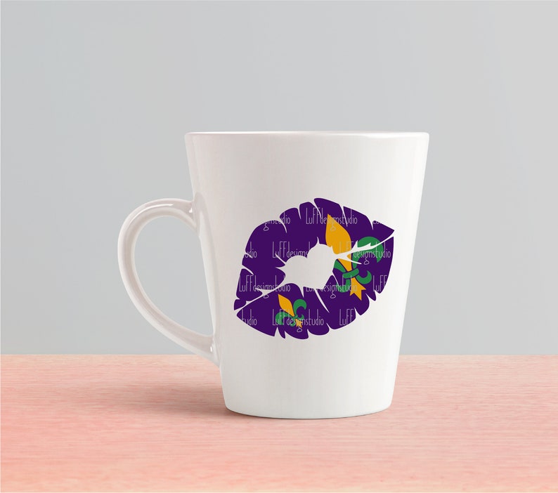 May include: White ceramic mug with a purple and gold lip print featuring a fleur-de-lis design. The mug has a handle on the right side.