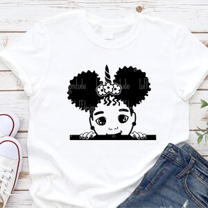 May include: A white t-shirt featuring a black and white illustration of a young girl with curly hair, a unicorn horn, and a flower headband peeking over a white bar.