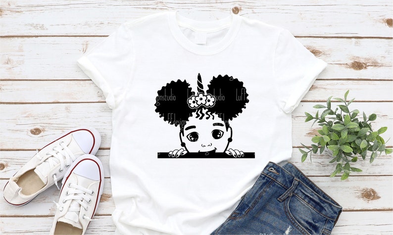 May include: A white t-shirt featuring a black and white illustration of a young girl with curly hair, a unicorn horn, and a flower headband peeking over a white bar.