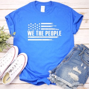 Op de afbeelding: Een helderblauwe t-shirt met de witte tekst "WE THE PEOPLE" en een gestileerd Amerikaans vlagontwerp. De afbeelding bevat een spijkerbroekshort en witte canvas sneakers. Het t-shirt is opgevouwen en de sneakers liggen op een licht houten oppervlak.