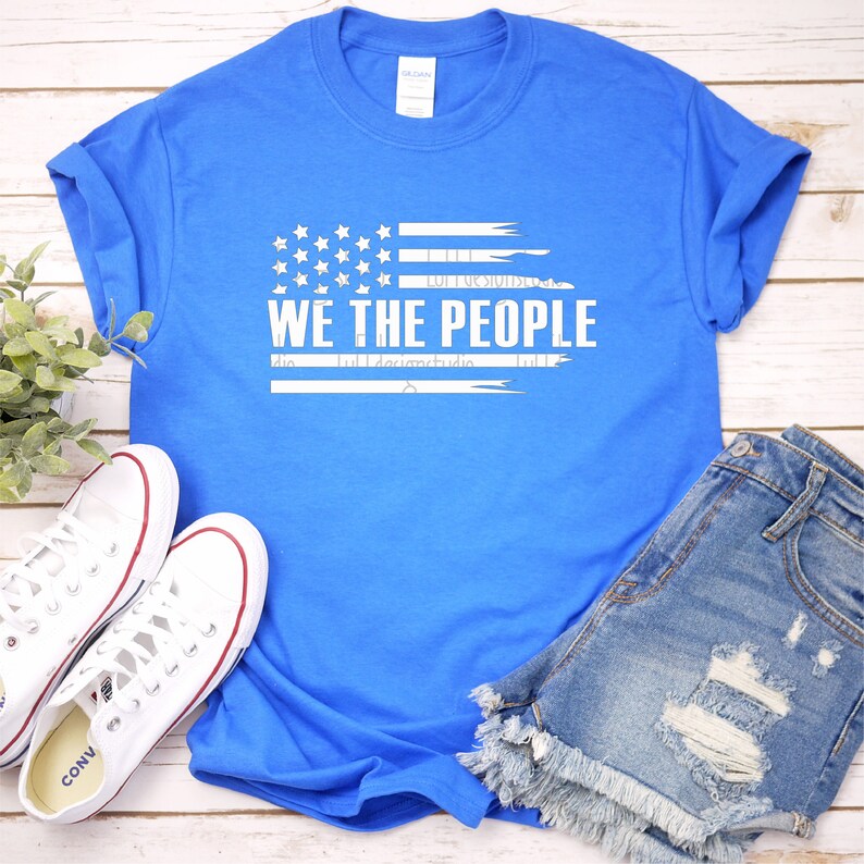 Op de afbeelding: Een helderblauwe t-shirt met de witte tekst "WE THE PEOPLE" en een gestileerd Amerikaans vlagontwerp. De afbeelding bevat een spijkerbroekshort en witte canvas sneakers. Het t-shirt is opgevouwen en de sneakers liggen op een licht houten oppervlak.
