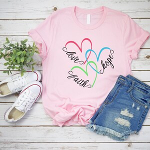 May include: Pink t-shirt with a design of three intertwined hearts in red, blue, and green. The words "love", "faith", and "hope" are written around the hearts.