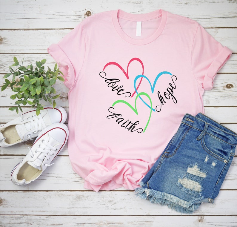 May include: Pink t-shirt with a design of three intertwined hearts in red, blue, and green. The words "love", "faith", and "hope" are written around the hearts.