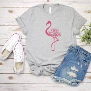 May include: A gray t-shirt with a pink flamingo graphic with pink flowers. The flamingo is standing on one leg. The t-shirt is laying on a white wooden surface with a pair of white sneakers and blue denim shorts.