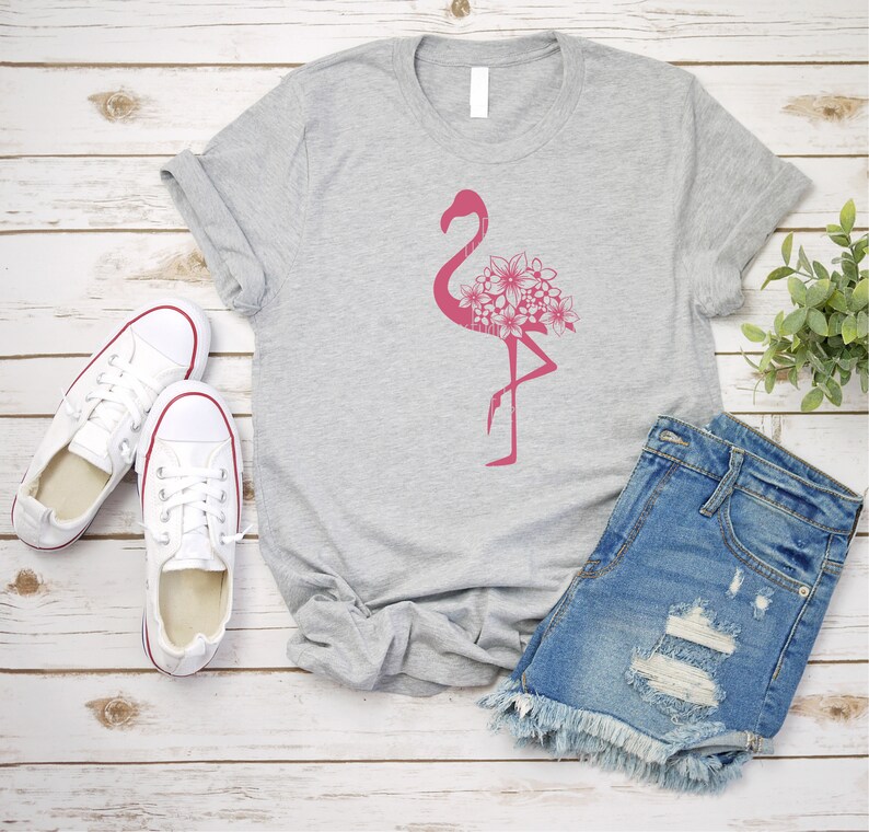 May include: A gray t-shirt with a pink flamingo graphic with pink flowers. The flamingo is standing on one leg. The t-shirt is laying on a white wooden surface with a pair of white sneakers and blue denim shorts.