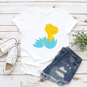 以下が含まれることがあります： 白いTシャツに、青い卵から孵化する黄色の恐竜が描かれています。恐竜は画像の右側を見ています。Tシャツは白い木製の表面に置かれ、白いスニーカーと青いデニムのショートパンツが置かれています。