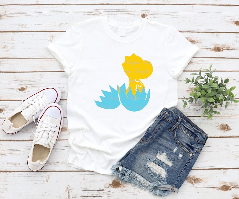 以下が含まれることがあります： 白いTシャツに、青い卵から孵化する黄色の恐竜が描かれています。恐竜は画像の右側を見ています。Tシャツは白い木製の表面に置かれ、白いスニーカーと青いデニムのショートパンツが置かれています。